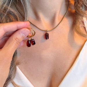 May include: A gold-toned chain necklace featuring a single rectangular deep red pendant, paired with matching hoop earrings that each hold a similar rectangular deep red charm. The jewelry set displays a consistent metallic finish and dark red stones.