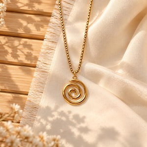 May include: A gold-toned necklace featuring a circular pendant with a raised spiral design. The pendant hangs from a delicate box chain. The metal has a polished, reflective finish that catches the light, creating a bright and metallic appearance.