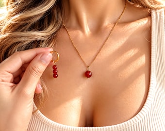 May include: A gold-toned chain necklace featuring a single round red bead pendant, paired with a matching gold-toned hoop earring that displays three smaller round red beads hanging vertically from the bottom of the hoop.
