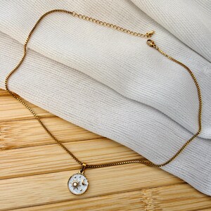 May include: A gold-toned box chain necklace featuring a circular pendant. The pendant has a white enamel background with a gold sun and crescent moon design, accented by small gold stars and a central clear stone set in the middle of the sun.