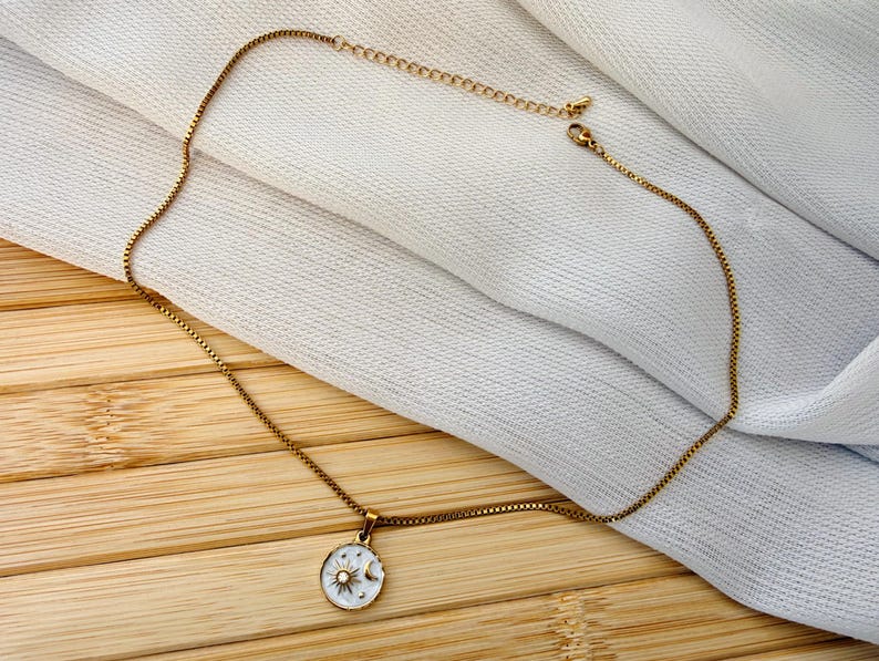 May include: A gold-toned box chain necklace featuring a circular pendant. The pendant has a white enamel background with a gold sun and crescent moon design, accented by small gold stars and a central clear stone set in the middle of the sun.