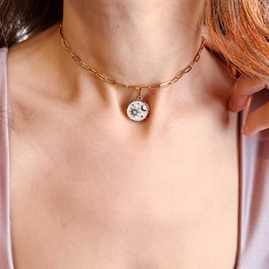 May include: A gold-toned chain necklace featuring a round pendant with a white enamel inlay. The pendant displays a black graphic design of a sun, a crescent moon, and small stars. The necklace is constructed with elongated oval links.