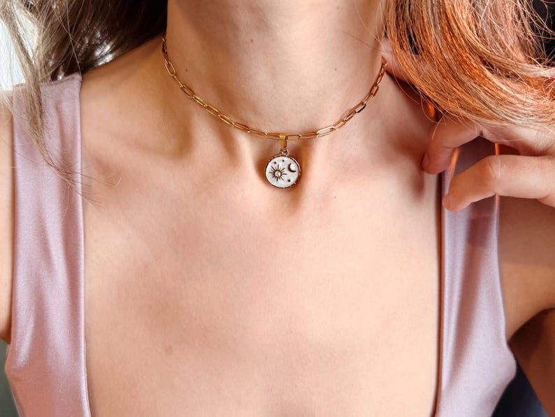 May include: A gold-toned chain necklace featuring a round pendant with a white enamel inlay. The pendant displays a black graphic design of a sun, a crescent moon, and small stars. The necklace is constructed with elongated oval links.