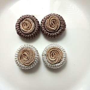 May include: Four circular buttons featuring a central spiral design in metallic gold thread. Two buttons have a dark brown braided border, while the other two buttons feature a silver braided border, all arranged in a two-by-two grid pattern.
