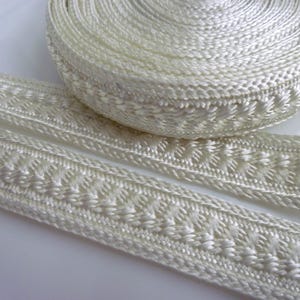 May include: Cream-coloured decorative trim with a woven design and a repeating pattern. A roll of the trim is visible, alongside two lengths of the trim laid out. Suitable for craft and sewing projects.