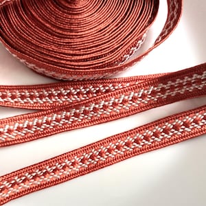 May include: A spool of rust-coloured woven ribbon featuring a repeating white geometric pattern along the centre. The ribbon has a smooth, slightly shiny texture and is coiled into a tight roll with a loose end extending outwards.