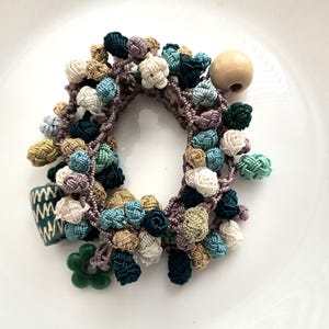 May include: A handmade hair tie featuring a macrame design with various colored knots in shades of green, beige, blue, and white. A wooden bead and decorative green flower and ceramic piece add detail to this unique hair accessory.