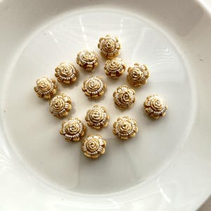 以下が含まれることがあります： 白地の表面に、金色の花型の小さな装飾品が並んでいます。各装飾品は、白い中心部と、花の中心に渦巻き模様のあるテクスチャ加工が施されています。装飾品は、花のデザインが特徴です。
