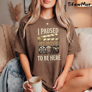 May include: Brown t-shirt with the text "I PAUSED MY MOVIE TO BE HERE" and a graphic of a film clapperboard, film reel, popcorn, and a film projector. The shirt is a relaxed fit.