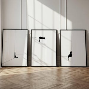 May include: Three framed art prints featuring minimalist black cat silhouettes on white backgrounds. Each print has a black frame and a simple line design. The prints are displayed against a white wall with a wooden floor.