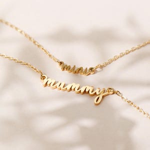May include: Two gold necklaces with the words "mummy" and "mine" hanging on a white surface.