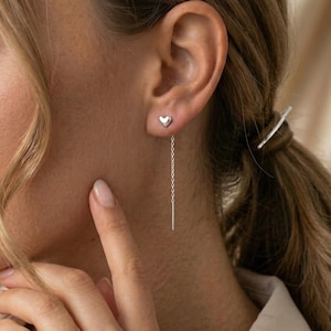 May include: A pair of silver earrings. One earring is a small heart stud, and the other is a chain with a vertical bar. The earrings are worn on the ear of a person with light brown hair.