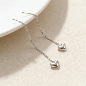 May include: A pair of silver-colored drop earrings. Each earring features a small, heart-shaped charm suspended from a delicate chain. The earrings are displayed on a white surface against a neutral background.
