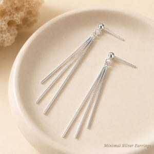May include: A pair of silver-toned earrings featuring a small ball stud post connected to three thin, dangling metallic bars of varying lengths. The earrings are displayed on a smooth, light-colored surface with the text Minimal Silver Earrings.