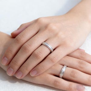 Op de afbeelding: Twee zilveren ringen met een band van kleine, heldere stenen. De ringen bevinden zich op twee handen, waarbij de ene hand op de andere rust. Dit zijn waarschijnlijk trouw- of verlovingsringen.