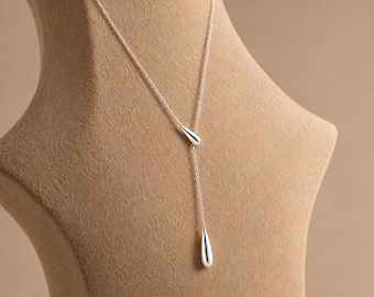 Peut inclure: Un collier lariat en argent composé d'une chaîne fine et de deux pendentifs polis en forme de goutte. Un pendentif est situé à la jonction centrale, tandis que le second est suspendu au bout de la chaîne. Le texte Minimal Silver Drop Necklace est visible.