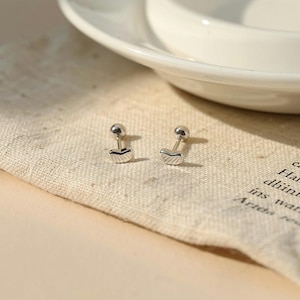 May include: Two silver stud earrings with a heart-shaped design. Each earring features a small, round ball at the top and a curved, heart-like shape below. The earrings are displayed on a beige fabric.
