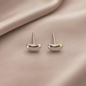 May include: A pair of stud earrings featuring a smooth, curved, bean-shaped design. Each earring is crafted with a polished silver-tone base and a contrasting gold-tone accent along the top edge, complete with a standard post back for secure wear.