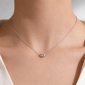 Può includere: Una collana d'argento con un piccolo pendente a forma di fagiolo. Il pendente è liscio e riflettente, sospeso a una catena sottile. La collana è mostrata su uno sfondo neutro, evidenziando il suo design minimalista.