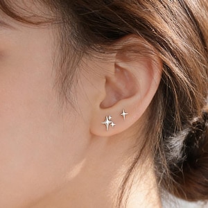 Puede incluir: Primer plano de una oreja adornada con dos pendientes de botón plateados en forma de estrella. Uno presenta un diseño de estrella de cuatro puntas, y el otro es una estrella simple de cuatro puntas más pequeña. Los pendientes están sobre la piel.