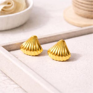 May include: Two gold-colored seashell-shaped stud earrings. The earrings have a ridged texture and a shiny finish. They are displayed on a light gray tray, with a blurred background that includes a white bowl and a wooden spool.