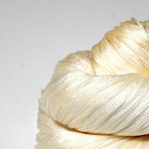 May include: A close-up of a spool of ivory satin ribbon. The ribbon is tightly wound and has a subtle sheen.