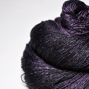 May include: A skein of dark purple yarn with a distinct metallic sheen and a smooth, twisted texture. The deep violet fibers catch the light, creating highlights across the coiled surface of the yarn, which is wound into a compact, rounded shape.