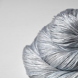 May include: A close-up of a skein of silver-gray yarn. The yarn is tightly wound, with visible strands creating a textured surface. The color is a blend of silver and gray, giving it a metallic sheen. Ideal for knitting or crochet projects.