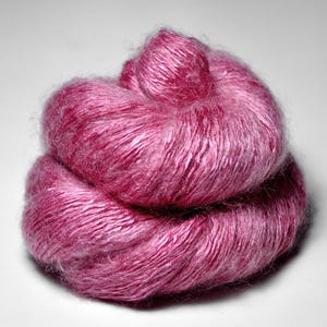 Puede incluir: Una madeja de hilo rosa, bien enrollada y texturizada. El hilo parece suave y esponjoso, con un ligero brillo. El color es un rosa medio vibrante, adecuado para proyectos de tejido o ganchillo.
