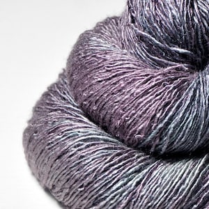May include: A close-up shot of a ball of yarn in shades of purple, grey, and teal. The yarn is tightly wound, with visible strands creating a textured surface. The colour transitions create a subtle gradient effect, suitable for knitting or crochet projects.