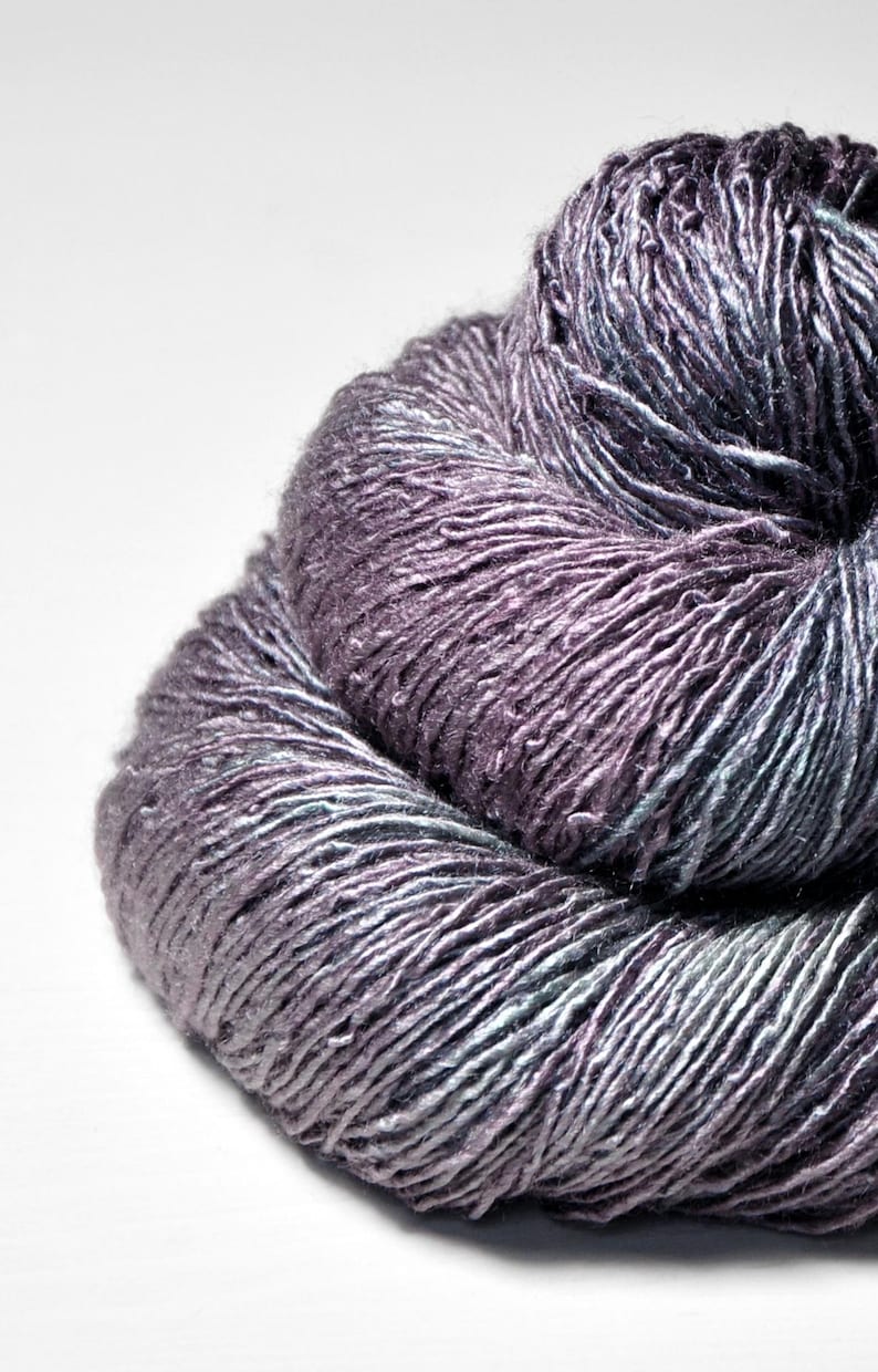 May include: A close-up shot of a ball of yarn in shades of purple, grey, and teal. The yarn is tightly wound, with visible strands creating a textured surface. The colour transitions create a subtle gradient effect, suitable for knitting or crochet projects.