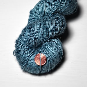 May include: Close-up of a skein of teal-colored yarn. The yarn is a deep, rich teal with subtle variations in color. A penny is placed next to the yarn for size comparison. The yarn appears soft and textured, suitable for knitting or crochet projects.
