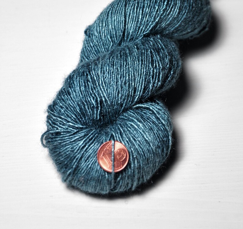 May include: Close-up of a skein of teal-colored yarn. The yarn is a deep, rich teal with subtle variations in color. A penny is placed next to the yarn for size comparison. The yarn appears soft and textured, suitable for knitting or crochet projects.