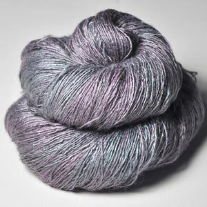 May include: A ball of yarn in a blend of grey, purple, and teal hues. The yarn is tightly wound, with visible strands creating a textured appearance. The colour shifts give the yarn an iridescent quality, suitable for knitting or crochet projects.