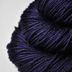 May include: Close-up of a skein of dark indigo yarn. The yarn is tightly wound, showing individual strands with a textured appearance. The color is a deep, rich blue with subtle variations in tone, suggesting a high-quality material.