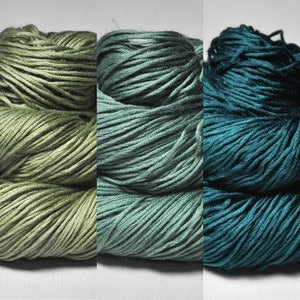 May include: Five skeins of yarn in a gradient of green and teal hues. The yarn is tightly wound, with visible strands creating a textured appearance. The colors range from a light, muted green to a deep teal.