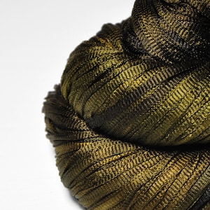 May include: Close-up of a tightly wound, textured fabric in shades of olive green and dark brown. The material appears to be woven with a ribbed pattern, creating a layered, dimensional effect. The fabric is bunched together, suggesting a scarf or other textile.