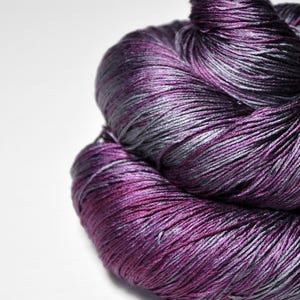 May include: A skein of yarn featuring a variegated color pattern of deep magenta, dark purple, and cool slate gray. The fibers have a smooth, lustrous sheen, with the strands tightly twisted together in a continuous, soft, and rounded bundle.