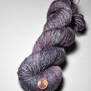 May include: A skein of hand-dyed yarn in shades of purple, grey, and silver. The yarn is twisted into a compact bundle, with a penny placed on the front for scale. The yarn appears soft and textured, suitable for knitting or crochet projects.