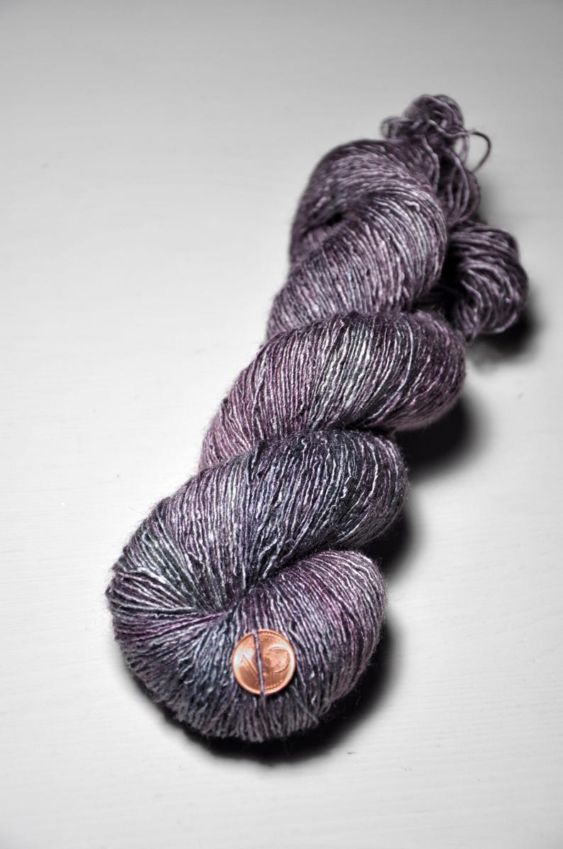 May include: A skein of hand-dyed yarn in shades of purple, grey, and silver. The yarn is twisted into a compact bundle, with a penny placed on the front for scale. The yarn appears soft and textured, suitable for knitting or crochet projects.