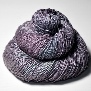 May include: A ball of yarn in shades of purple, grey, and teal. The yarn is tightly wound, showing the texture of the fibres. The colour transitions create a subtle gradient effect, with the darker hues concentrated in the centre.