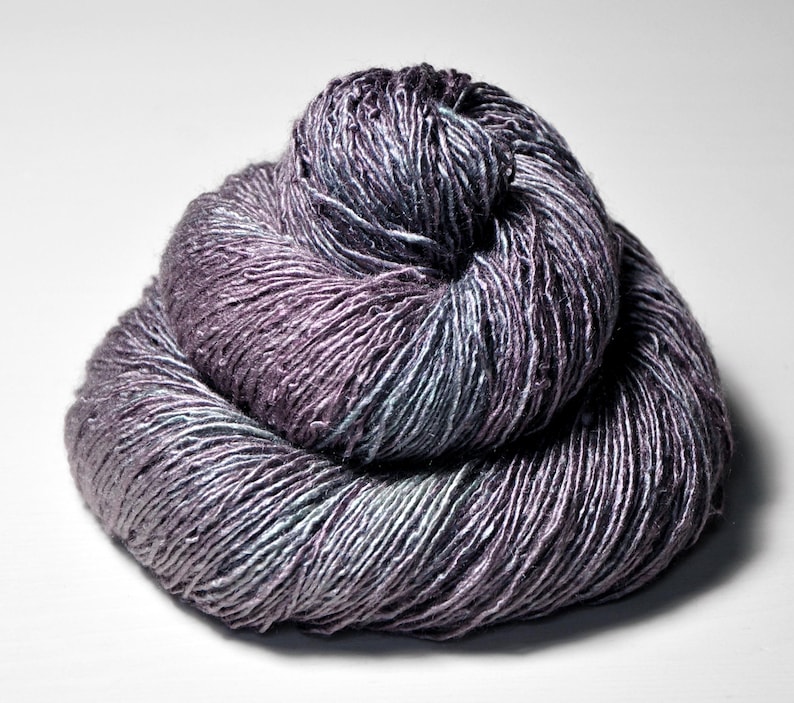 May include: A ball of yarn in shades of purple, grey, and teal. The yarn is tightly wound, showing the texture of the fibres. The colour transitions create a subtle gradient effect, with the darker hues concentrated in the centre.