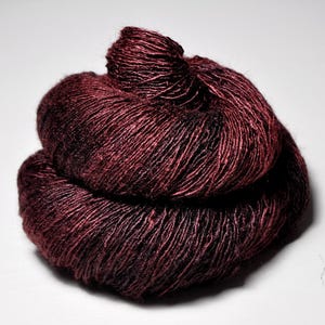 May include: A close-up of a skein of burgundy-colored yarn. The yarn is tightly wound, with visible strands and a slightly textured appearance. The color is a deep, rich shade of red with hints of black.