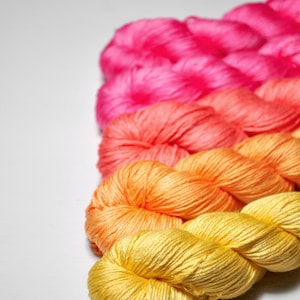 May include: A selection of colourful yarn skeins in shades of pink, coral, orange, and yellow. The yarn is arranged in a row, with each colour transitioning to the next. The skeins are tightly wound and have a soft, textured appearance.