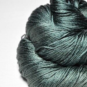 May include: A skein of dark teal yarn with a distinct sheen and smooth texture. The fibers are tightly twisted together, creating a deep, reflective surface that transitions between shades of forest green and blue in the light.