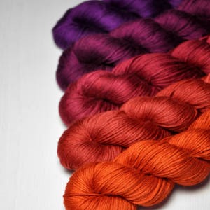 Könnte beinhalten: Eine Sammlung farbenfroher Garnknäuel in einem Farbverlauf. Die Farben reichen von tiefem Lila bis zu leuchtendem Orange. Das Garn ist diagonal vor einem weißen Hintergrund angeordnet, wodurch die Textur und Farbvariationen hervorgehoben werden.