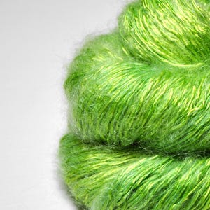 May include: Close-up of a vibrant green yarn skein with streaks of yellow. The fluffy texture of the yarn is visible against a white background. This yarn is likely used for knitting or crocheting projects.