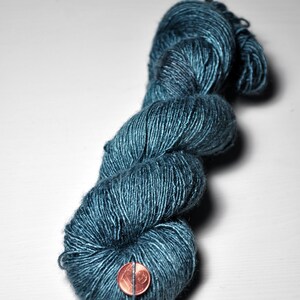 May include: A skein of teal-colored yarn, tightly wound and textured. The yarn is a deep, saturated blue-green hue. A penny is placed on the yarn for scale. Ideal for knitting, crocheting, or other fiber arts projects.