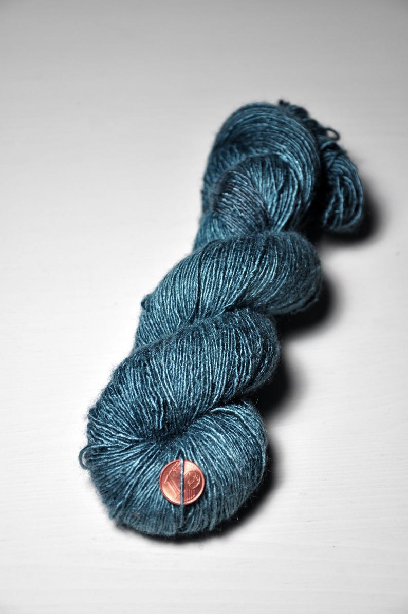 May include: A skein of teal-colored yarn, tightly wound and textured. The yarn is a deep, saturated blue-green hue. A penny is placed on the yarn for scale. Ideal for knitting, crocheting, or other fiber arts projects.