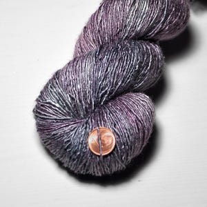 May include: A ball of hand-dyed yarn in shades of purple, grey, and silver. The yarn has a variegated colour scheme with a subtle sheen. A penny is placed on the yarn for size reference. Suitable for knitting or crochet projects.